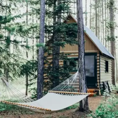 A cozy log cabin nestled in a dense forest, with a hammock strung between two trees in the foreground. The scene conveys tranquility and solitude.