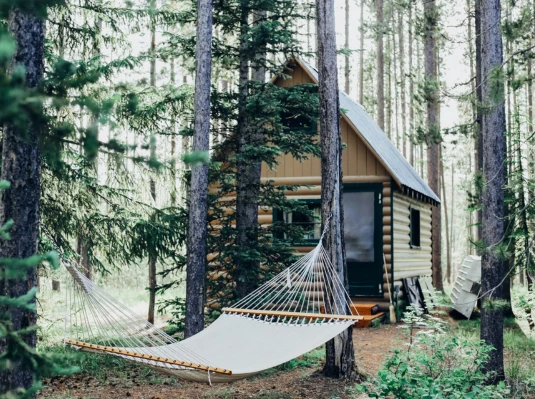 A cozy log cabin nestled in a dense forest, with a hammock strung between two trees in the foreground. The scene conveys tranquility and solitude.