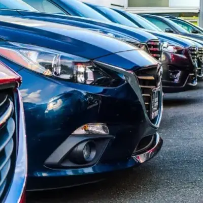 Close-up of a row of parked cars with shiny front grilles and headlights reflecting light, conveying a sleek and modern tone. Asphalt road visible.