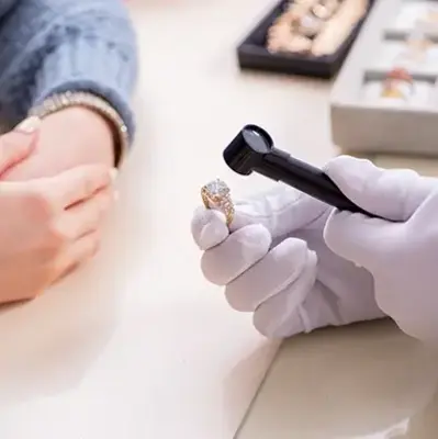 A jeweler in white gloves examines a diamond ring with a magnifying glass. A person with folded hands waits nearby. Rings are displayed in the background.