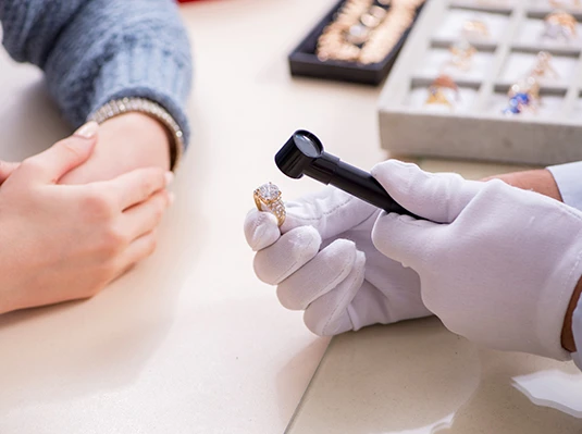 A jeweler in white gloves examines a diamond ring with a magnifying glass. A person with folded hands waits nearby. Rings are displayed in the background.