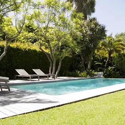 A serene backyard scene with a sparkling pool, surrounded by lush green trees and bushes. Two empty loungers sit on a sunlit deck, evoking tranquility.