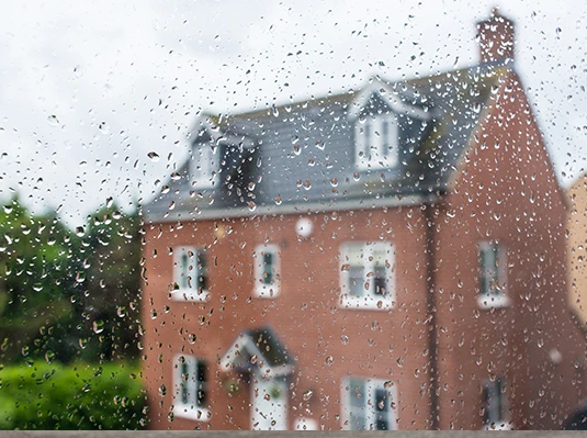 Raindrops on a window blur the view of a red brick house with a gabled roof and white-framed windows, conveying a calm, rainy day atmosphere.