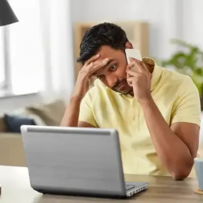 A man in a yellow shirt looks stressed while talking on the phone at a desk. A laptop is open in front of him, and a plant is in the background.