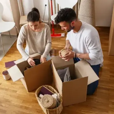 A couple is packing cardboard boxes in a room with wooden floors. Nearby are a suitcase, chairs, and a basket. They appear focused and busy.