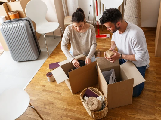 A couple is packing cardboard boxes in a room with wooden floors. Nearby are a suitcase, chairs, and a basket. They appear focused and busy.