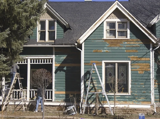 Two workers on ladders paint an old, weathered green house with peeling paint. The scene conveys renovation and renewal under clear blue skies.