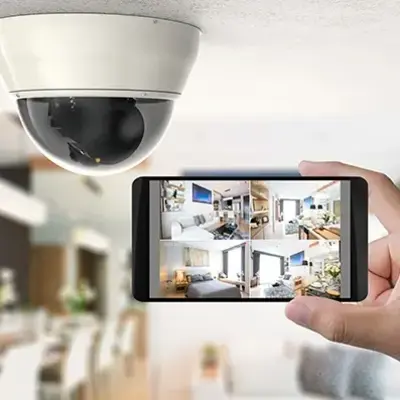 A hand holds a smartphone showing multiple home security camera feeds. A dome-shaped security camera is mounted on the ceiling in a bright, modern living space.