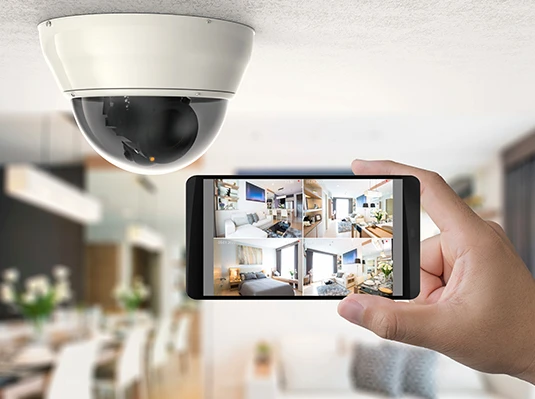 A hand holds a smartphone showing multiple home security camera feeds. A dome-shaped security camera is mounted on the ceiling in a bright, modern living space.