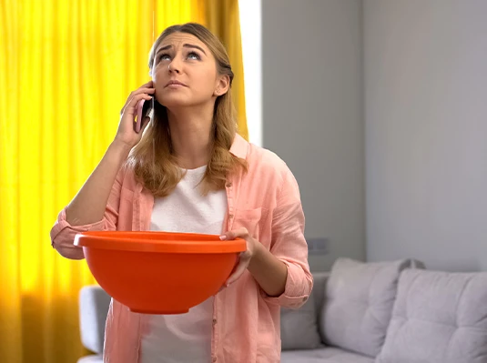 A worried woman holds a large orange bowl under a ceiling leak while talking on the phone. She stands in a living room with yellow curtains and a gray sofa.