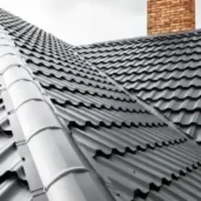 A modern house roof with sleek, dark gray metal tiles, and a red brick chimney against a cloudy sky, conveying a sense of durability and contemporary design.