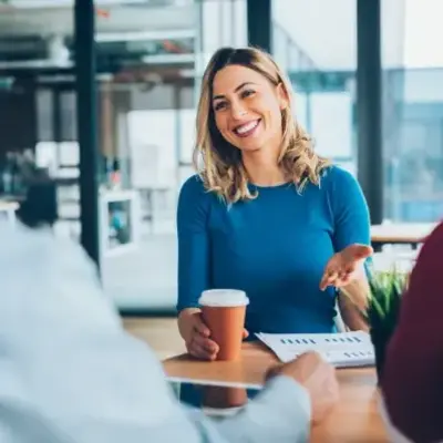 A woman in a blue shirt, smiling, holds a coffee cup during a meeting. Two people, blurred, converse with her in a bright office setting.