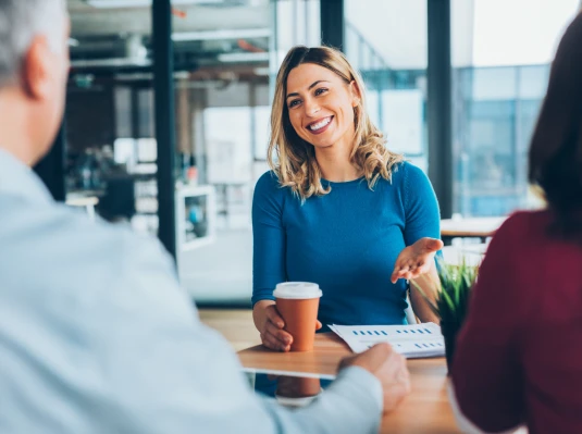 A woman in a blue shirt, smiling, holds a coffee cup during a meeting. Two people, blurred, converse with her in a bright office setting.