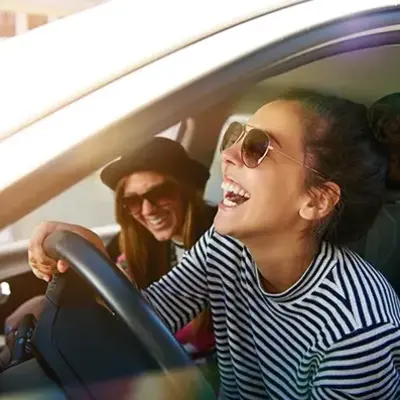 Two women, wearing sunglasses, laugh joyfully inside a car. One is in the driver's seat, gripping the wheel, conveying a tone of fun and happiness.