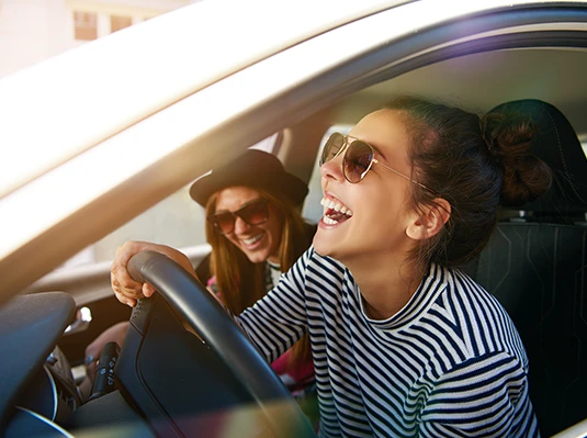 Two women, wearing sunglasses, laugh joyfully inside a car. One is in the driver's seat, gripping the wheel, conveying a tone of fun and happiness.