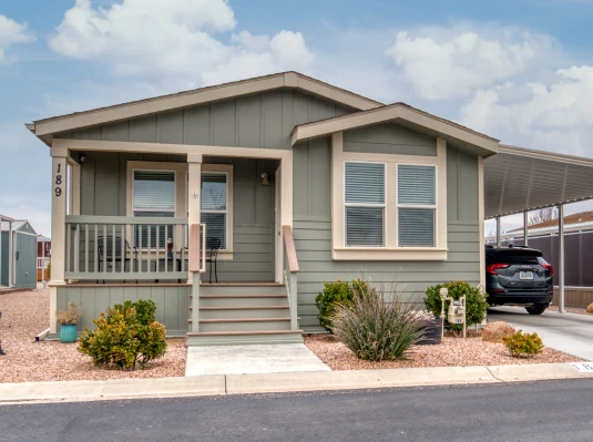 Small, green manufactured home with front porch and desert landscaping. Covered carport with black SUV. Sky is partly cloudy, creating a serene atmosphere.