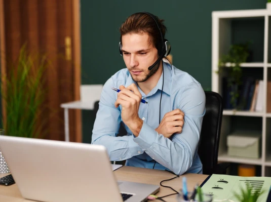 A man with a headset sits at a desk, focused on his laptop, engaged in a virtual meeting or task.
