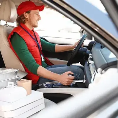 A man wearing a red cap and vest is driving a car, with food delivery items on the passenger seat. The scene conveys a sense of focus and purpose.