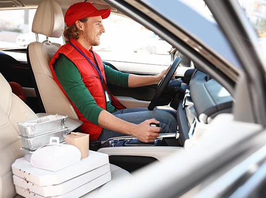 A man wearing a red cap and vest is driving a car, with food delivery items on the passenger seat. The scene conveys a sense of focus and purpose.