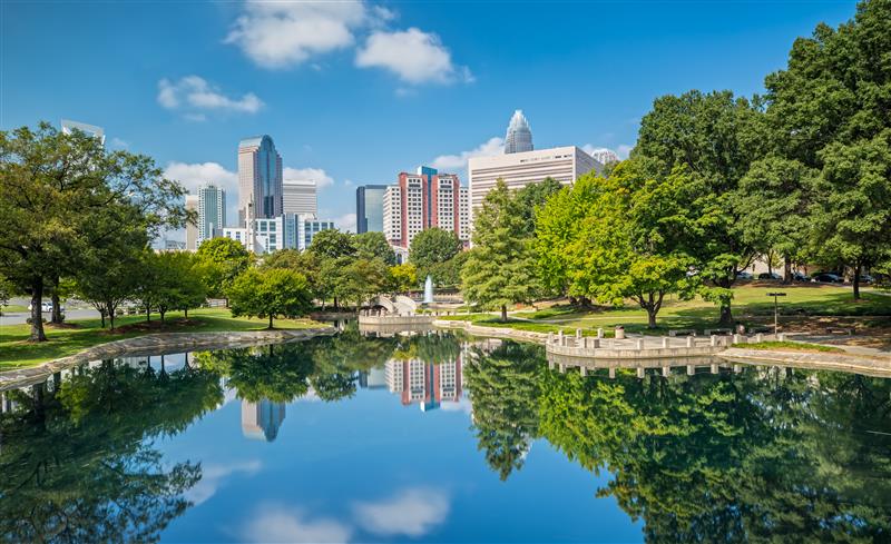Charlotte, North Carolina skyline reflecting in a serene pond, surrounded by lush greenery and trees under a clear blue sky.